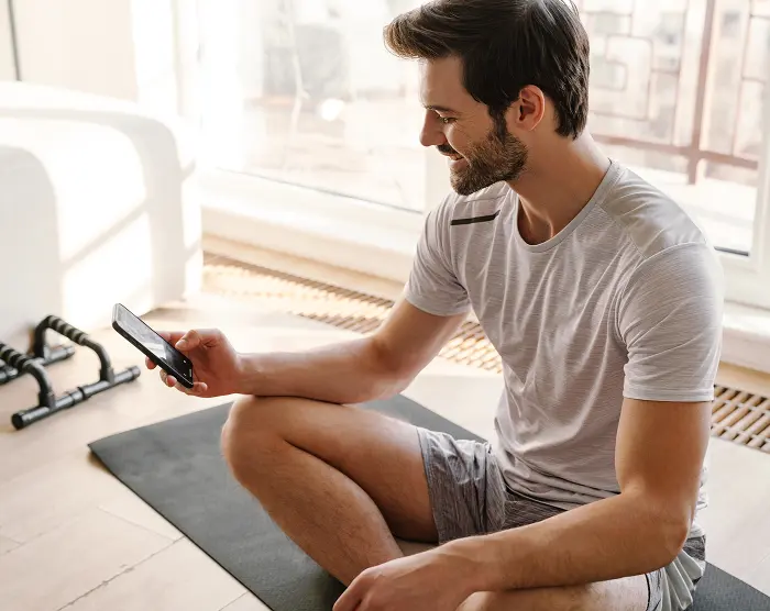 Man sitting on a workout mat at home, looking at his phone.