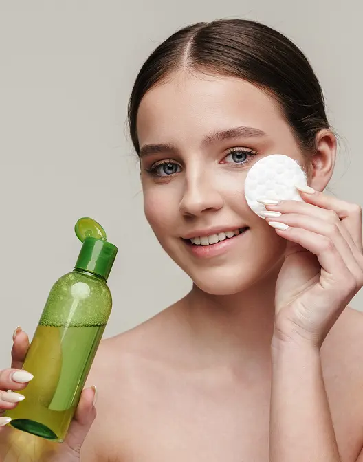 Young woman cleansing her face with a cotton pad while holding a green eco cosmetics bottle.