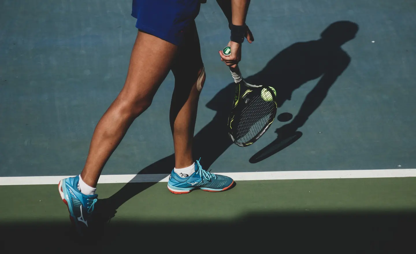 Tennis player's legs and racket on a sunlit hard court, with a ball resting on the strings.