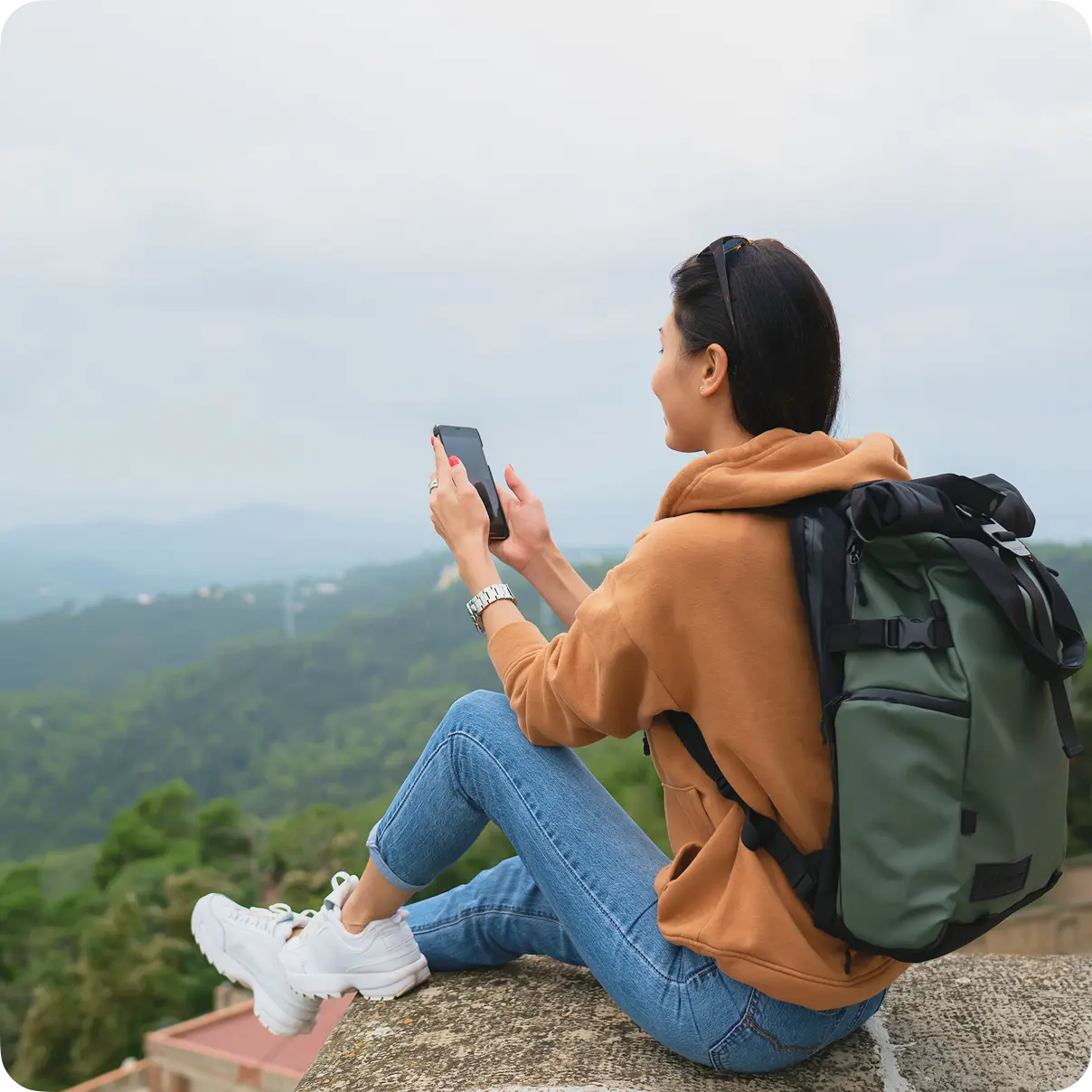 Traveler with a backpack sitting above a green valley and looking at a phone.