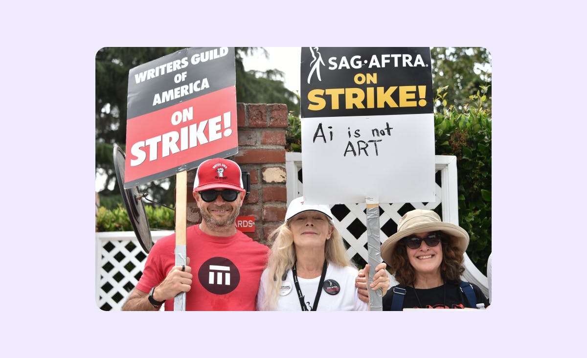 The photo features three protesters holding signs from SAG AFTRA and the Writers Guild of America. The man holds a sign from the writers’ guild that reads "On strike." The two women hold a sign from SAG AFTRA that reads "AI is not Art."