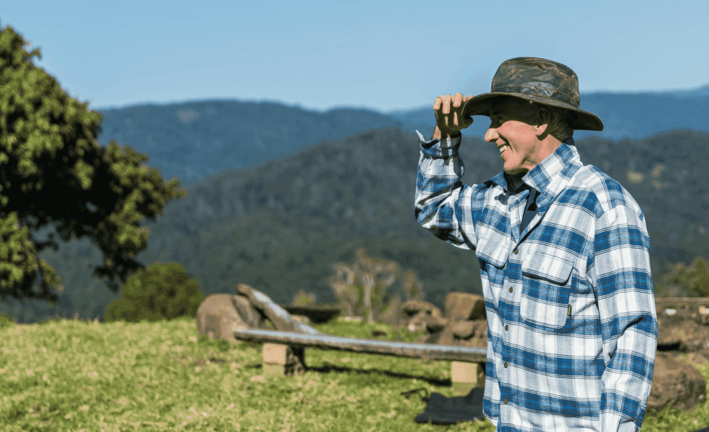 A photo of a smiling farmer in a hat and a plaid shirt in front of the green hills illustrating the purpose of the Noah mobile app for purchasing products directly from farmers