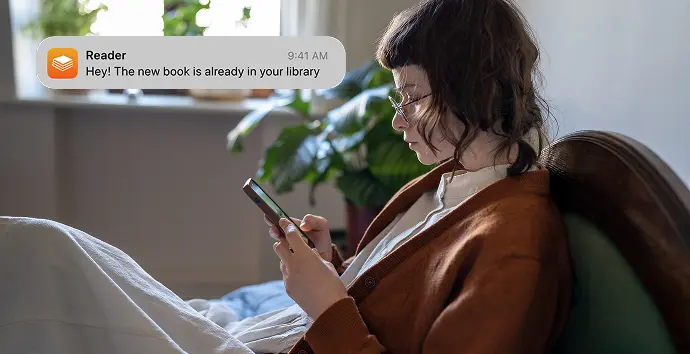 Woman reading on a phone in an armchair with a notification about a new book in her library.