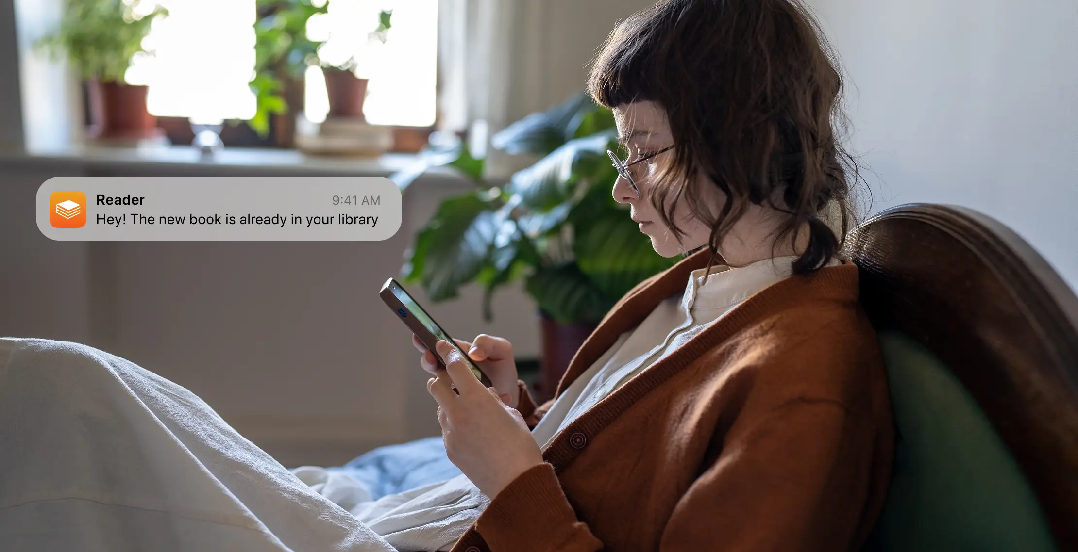 Woman reading on a phone in an armchair with a notification about a new book in her library.