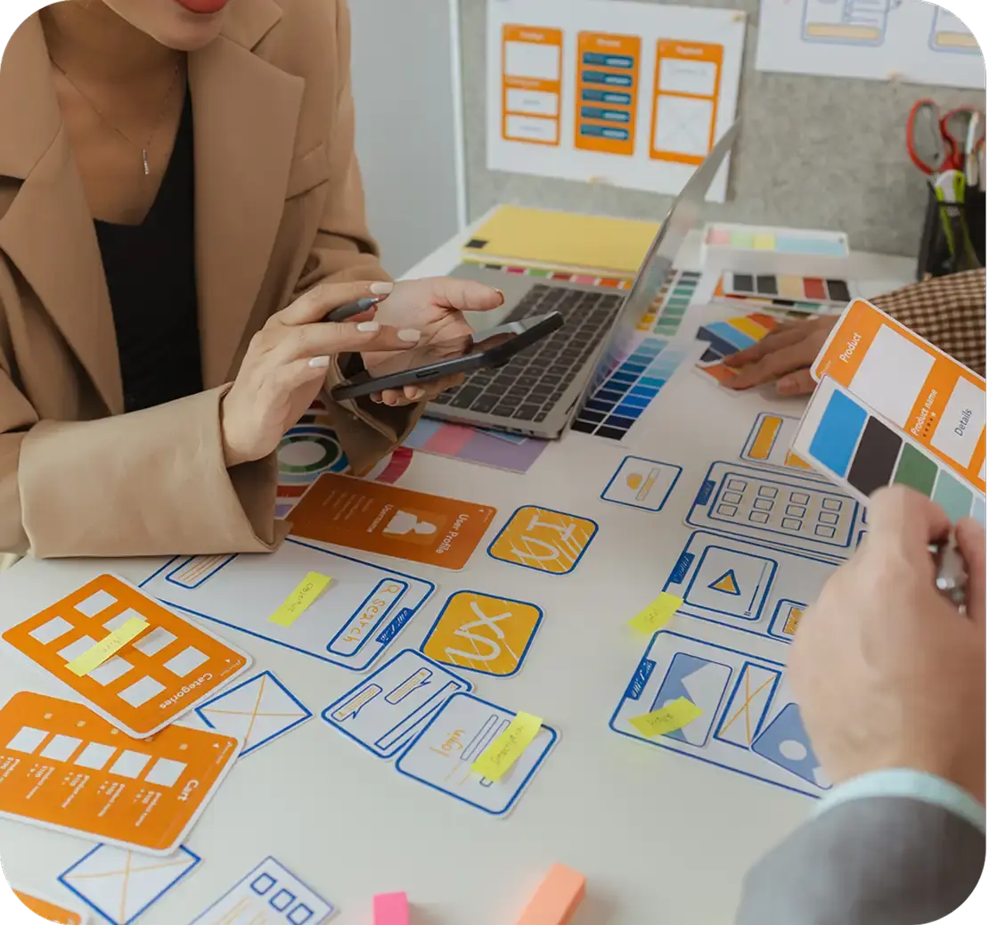 Two designers at a table covered with paper app screen mockups, sticky notes, a laptop, and color swatches.