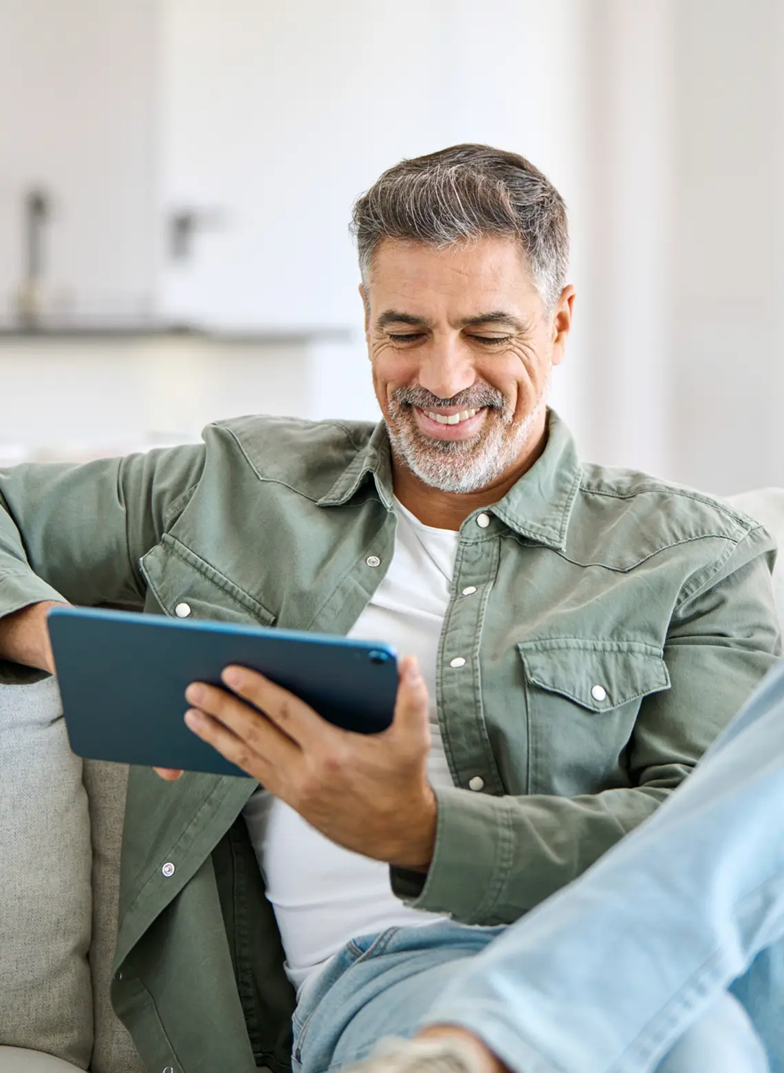 Smiling man sitting on a couch and reading on a tablet.
