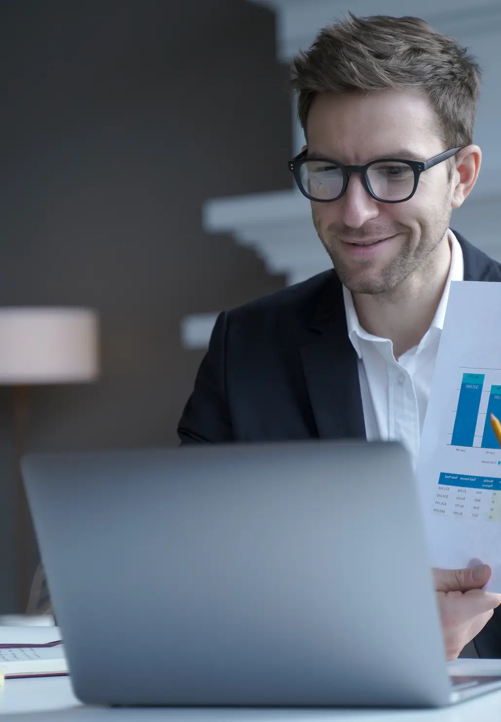Man in glasses at a laptop holding a printed report with bar charts.